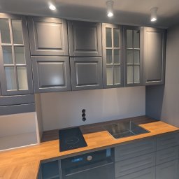 MJM PLUS Mateusz Jamioł - Szara zabudowa kuchenna z drewnianym blatem, widoczne szafki górne z przeszkleniami, płyta indukcyjna i zlew. Minimalistyczny design, nowoczesne oświetlenie.