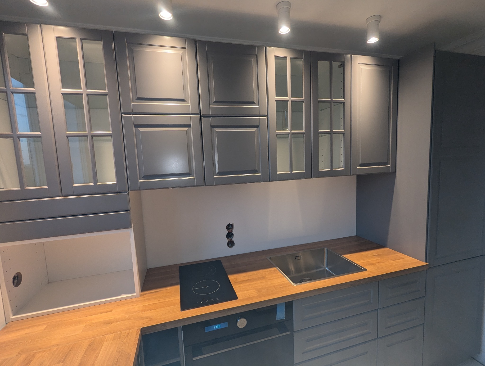 Szara zabudowa kuchenna z drewnianym blatem, widoczne szafki górne z przeszkleniami, płyta indukcyjna i zlew. Minimalistyczny design, nowoczesne oświetlenie.