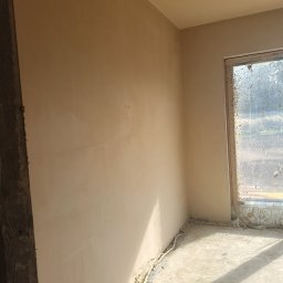 KRZYZ KACPER KRZYŻ - Świeżo otynkowane wnętrze z oknem, widoczne przewody przy podłodze. Ściany gładkie, w jasnym odcieniu beżu. Okno brudne, z resztkami zaprawy. Fragment drewnianej ramy w lewym rogu.