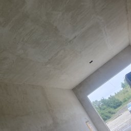 KRZYZ KACPER KRZYŻ - Surowe, jasne wnętrze po tynkowaniu maszynowym. Widoczne ślady po zacieraniu, sufit i ściany w odcieniach szarości. W tle okno z widokiem na zieleń i zaparkowany samochód.