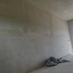 KRZYZ KACPER KRZYŻ - Wnętrze po nałożeniu szarego tynku na ściany i sufit. Widoczne kable wystające ze ściany i plandeka na podłodze. Okno z widokiem na zieleń w tle.