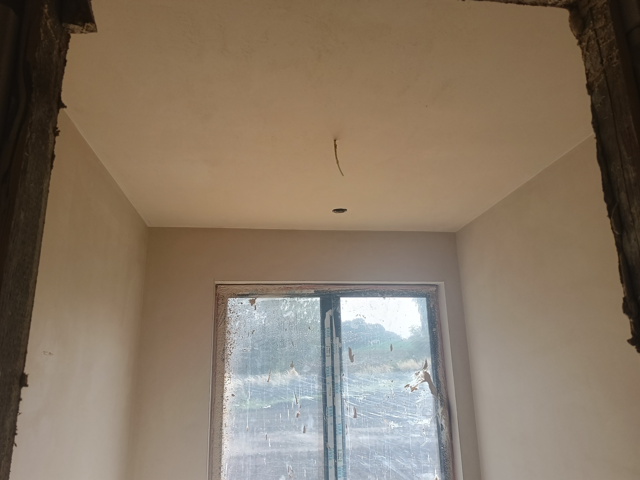 Wnętrze po nałożeniu gładzi gipsowej: ściany i sufit w jasnym odcieniu, okno zabezpieczone folią. Widoczne ślady po instalacji elektrycznej w suficie.