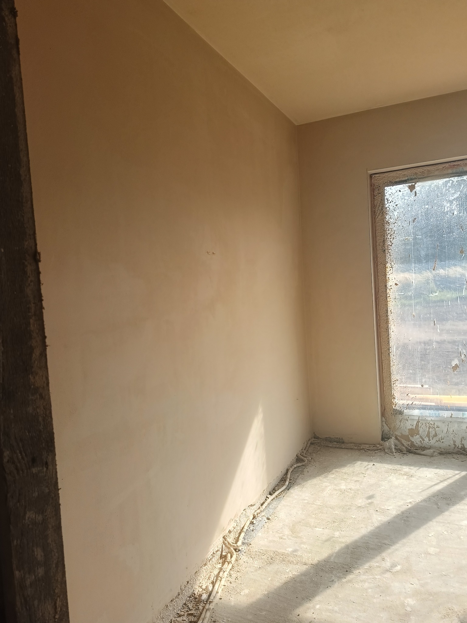 Świeżo otynkowane wnętrze z oknem, widoczne przewody przy podłodze. Ściany gładkie, w jasnym odcieniu beżu. Okno brudne, z resztkami zaprawy. Fragment drewnianej ramy w lewym rogu.