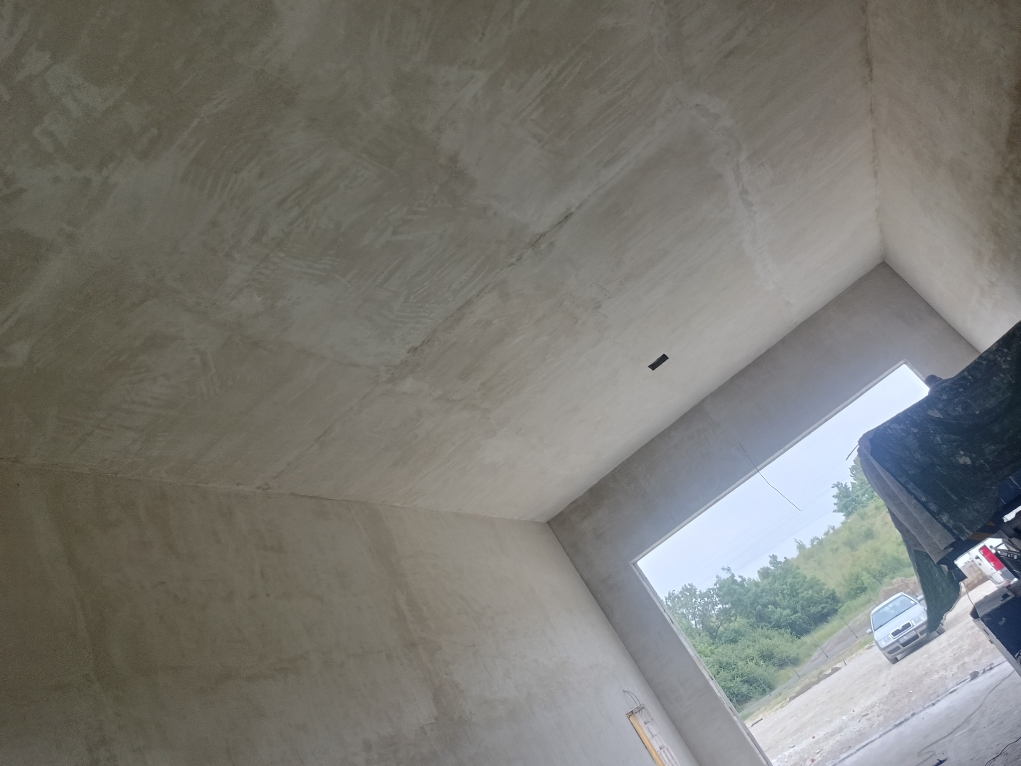 Surowe, jasne wnętrze po tynkowaniu maszynowym. Widoczne ślady po zacieraniu, sufit i ściany w odcieniach szarości. W tle okno z widokiem na zieleń i zaparkowany samochód.