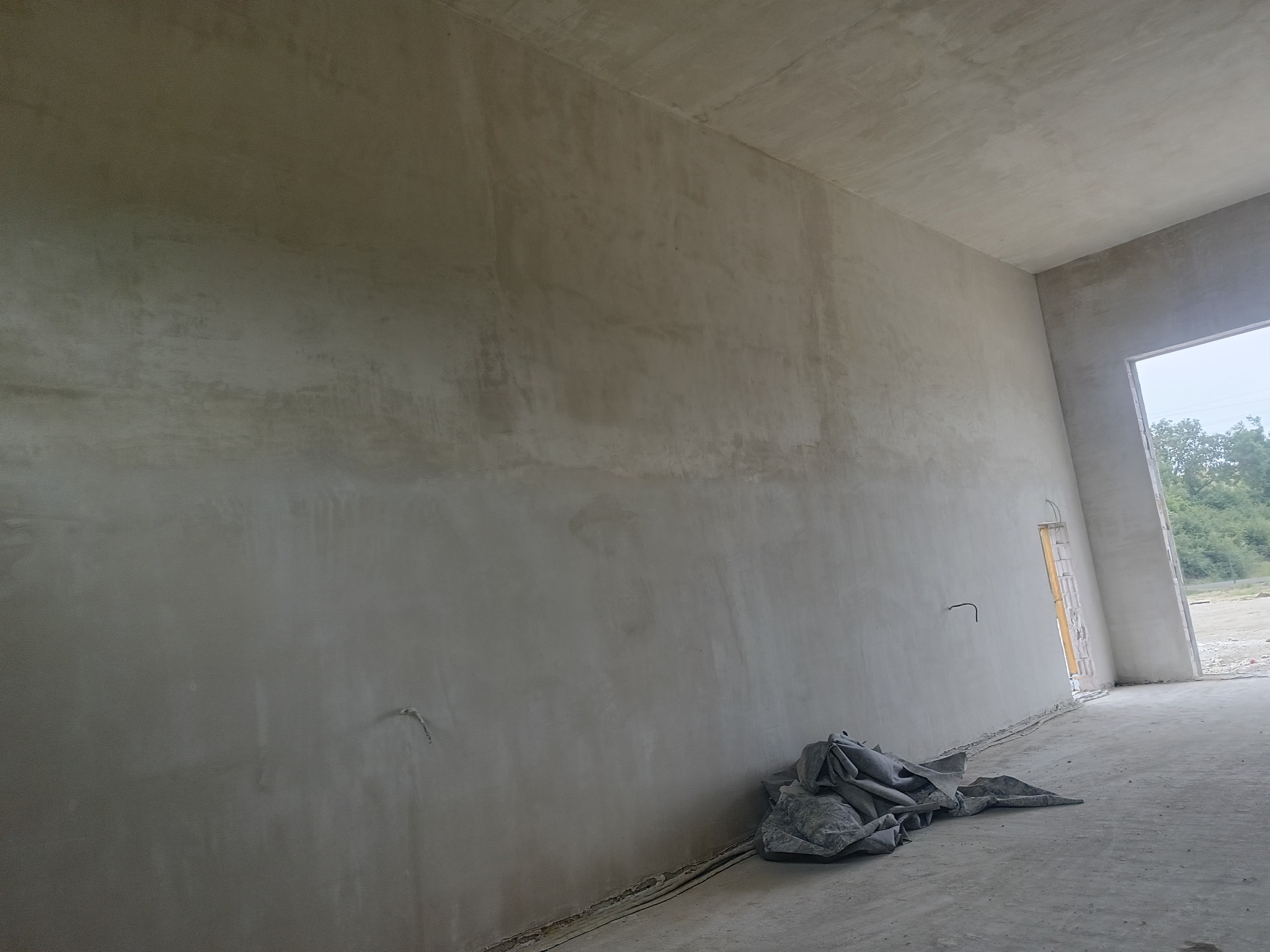 Wnętrze po nałożeniu szarego tynku na ściany i sufit. Widoczne kable wystające ze ściany i plandeka na podłodze. Okno z widokiem na zieleń w tle.