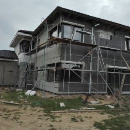 Dwukondygnacyjny dom w trakcie ocieplania, z widocznymi rusztowaniami i szarymi płytami izolacyjnymi. Częściowo otynkowana ściana boczna. Pochmurne niebo w tle.