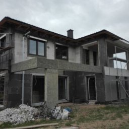 Dom w trakcie prac elewacyjnych: widoczne rusztowania, izolacja termiczna ścian i przygotowane materiały budowlane. Surowy stan, okna z roletami zewnętrznymi.