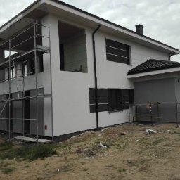 Dwupiętrowy dom w trakcie prac elewacyjnych. Część budynku z rusztowaniem, druga część z oknami i wykończoną elewacją w odcieniach bieli i szarości.