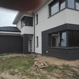 Nowoczesny dom z elewacją w odcieniach szarości i bieli, z widocznym fragmentem garażu i stertą drewna na trawniku. Minimalistyczny design, duże okna.