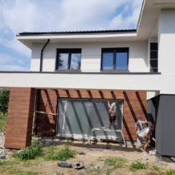 MMBUDMariuszMusinski - Montaż paneli elewacyjnych imitujących drewno na nowoczesnym domu z białymi ścianami i czarnymi oknami. Dwóch pracowników na drabinie i przy gruncie.