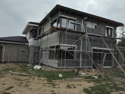 Dwukondygnacyjny dom w trakcie ocieplania, z widocznymi rusztowaniami i szarymi płytami izolacyjnymi. Częściowo otynkowana ściana boczna. Pochmurne niebo w tle.