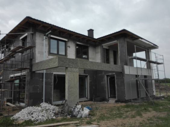 Dom w trakcie prac elewacyjnych: widoczne rusztowania, izolacja termiczna ścian i przygotowane materiały budowlane. Surowy stan, okna z roletami zewnętrznymi.