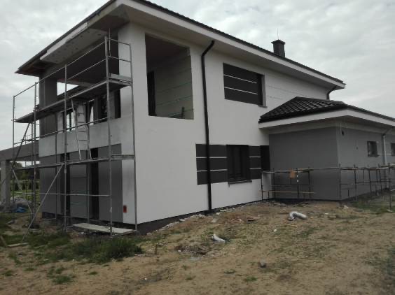 Dwupiętrowy dom w trakcie prac elewacyjnych. Część budynku z rusztowaniem, druga część z oknami i wykończoną elewacją w odcieniach bieli i szarości.