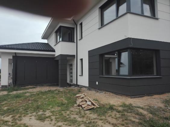 Nowoczesny dom z elewacją w odcieniach szarości i bieli, z widocznym fragmentem garażu i stertą drewna na trawniku. Minimalistyczny design, duże okna.