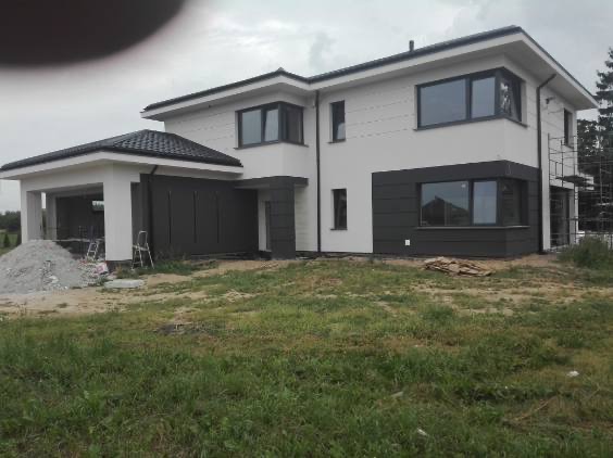 Nowoczesny dom jednorodzinny z białą elewacją i grafitowymi akcentami. Widoczne rusztowania sugerują prace wykończeniowe. Na pierwszym planie trawa i materiały budowlane.