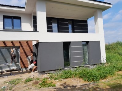 Montaż elewacji domu jednorodzinnego z wykorzystaniem paneli drewnopodobnych i grafitowych. Trzech pracowników w trakcie pracy.