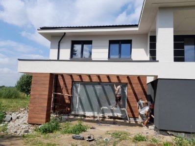 Montaż paneli elewacyjnych imitujących drewno na nowoczesnym domu z białymi ścianami i czarnymi oknami. Dwóch pracowników na drabinie i przy gruncie.