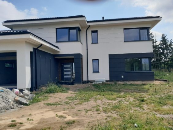 Nowoczesny dom jednorodzinny z biało-grafitową elewacją, widok z zewnątrz. Dwukondygnacyjny budynek z garażem, duże okna, minimalistyczny design. Teren wokół domu w trakcie zagospodarowania.