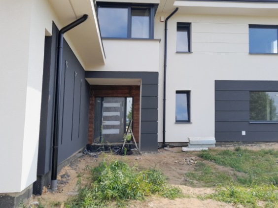 Nowoczesny dom z elewacją w odcieniach bieli i szarości, z widocznym wejściem i oknami. Uwagę zwraca kontrast kolorów i minimalistyczny design.