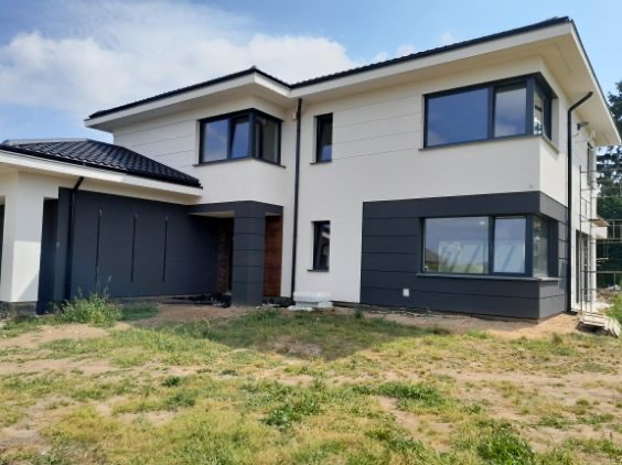 Dwukondygnacyjny dom z białą elewacją i grafitowymi panelami. Widoczne okna z ciemnymi ramami i fragment rusztowania. Błękitne niebo w tle.