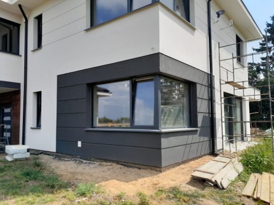 Nowoczesna elewacja domu jednorodzinnego z szarymi panelami i białym tynkiem. Widoczne okno narożne i fragment rusztowania. Estetyczne wykończenie.