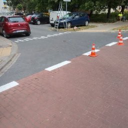 DROG-ZNAK SPÓŁKA Z OGRANICZONĄ ODPOWIEDZIALNOŚCIĄ - Oznakowane przejście dla pieszych z nowymi, białymi pasami i pomarańczowymi pachołkami. Widoczne samochody i osoba siedząca na ławce w tle.