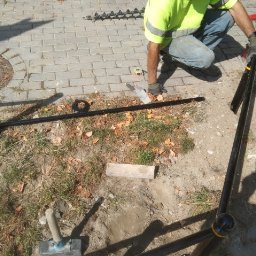 DROG-ZNAK SPÓŁKA Z OGRANICZONĄ ODPOWIEDZIALNOŚCIĄ - Montaż barierek ochronnych przy chodniku z kostki brukowej. Widoczny pracownik w odblaskowej kamizelce i narzędzia. Etap prac ziemnych przed betonowaniem.