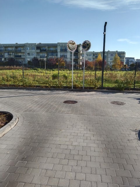 Ułożona kostka brukowa na drodze osiedlowej z widocznymi lustrami drogowymi i budynkami w tle, w słoneczny dzień. Widok z poziomu chodnika.