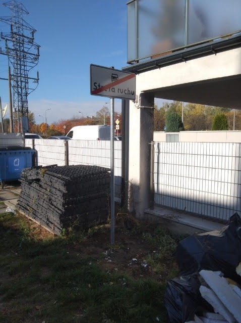 Stos czarnych płyt drogowych obok znaku 'Strefa ruchu' na tle białego ogrodzenia i słupa. Widoczne ślady prac budowlanych i elementy infrastruktury drogowej.