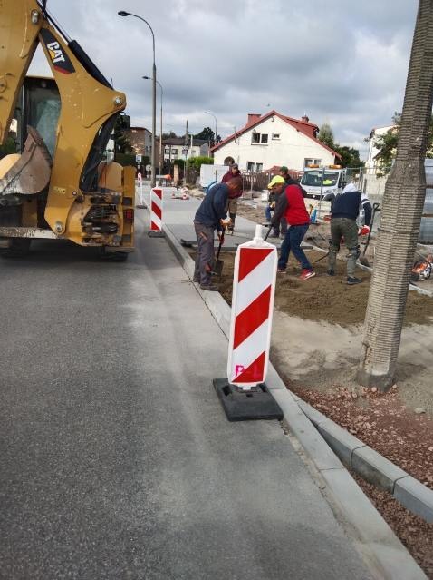 Ekipa robotników z łopatami i koparka CAT podczas budowy chodnika przy ulicy. Widoczne biało-czerwone słupki ostrzegawcze.