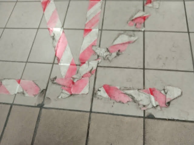Resztki czerwono-białej taśmy ostrzegawczej, częściowo odklejonej, leżą na szarych płytkach chodnikowych. Fragmenty taśmy tworzą nieuporządkowany wzór na powierzchni.
