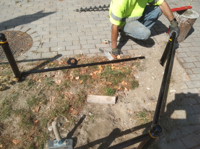 Montaż barierek ochronnych przy chodniku z kostki brukowej. Widoczny pracownik w odblaskowej kamizelce i narzędzia. Etap prac ziemnych przed betonowaniem.