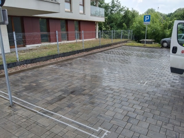 Ukończony parking z kostki brukowej z wyznaczonymi miejscami postojowymi, obok budynku mieszkalnego i znaku drogowego.