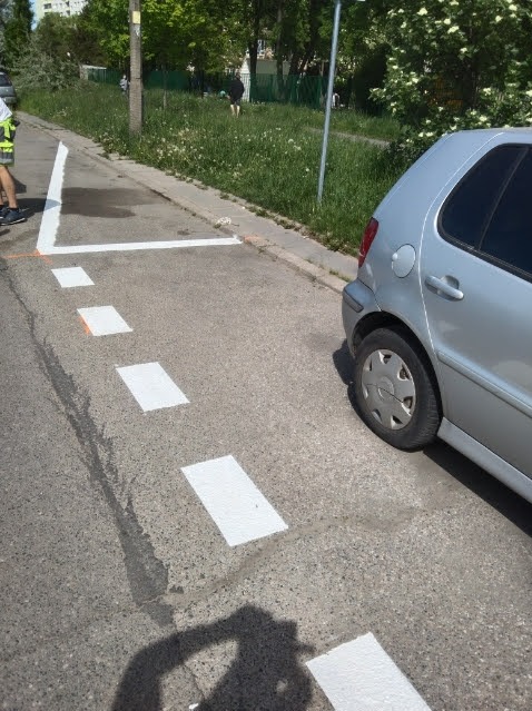 Oznakowane białą farbą miejsce parkingowe na asfalcie, częściowo zajęte przez srebrny samochód osobowy. Widoczny fragment chodnika i trawnika.