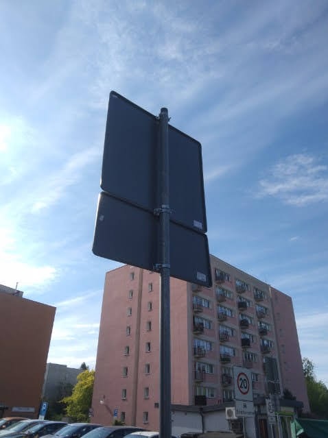 Drogowy znak pionowy na metalowym słupku, z budynkiem w tle i fragmentem parkingu. Widok z dołu, niebieskie niebo.