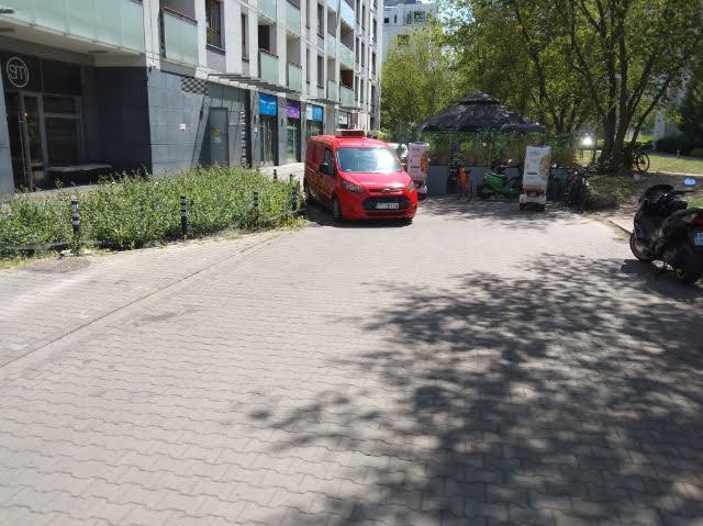 Czerwony samochód dostawczy zaparkowany na chodniku z kostki brukowej przed budynkiem. W tle zieleń i parasol. Cień drzewa na kostce. Słoneczny dzień.