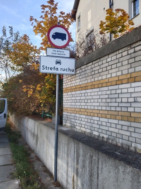 Znak drogowy 'Strefa ruchu' z zakazem wjazdu dla pojazdów ciężarowych, umieszczony na szarym słupku przy murze z cegły. Jesienne drzewa w tle.