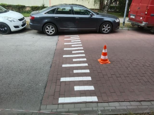 Oznakowanie poziome na drodze z kostki brukowej, z pomarańczowym pachołkiem. Widoczne samochody i fragment czerwonego busa w tle. Białe pasy tworzą wzór.