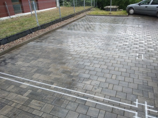 Wykończony parking z kostki brukowej z wyznaczonymi białymi liniami miejsc postojowych, widok z góry, częściowo widoczny samochód i zieleń w tle.