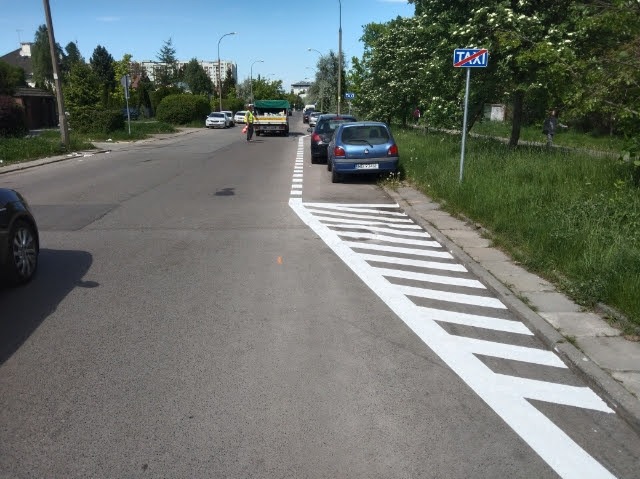 Świeżo wymalowane, białe pasy na asfalcie wyznaczają miejsce postojowe dla taksówek przy ulicy. W tle zaparkowane auta i zieleń drzew.
