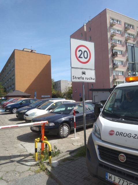 Znak drogowy 'Strefa ruchu' z ograniczeniem prędkości do 20 km/h na tle parkingu z samochodami i budynkami mieszkalnymi. Widoczny samochód z logo firmy.