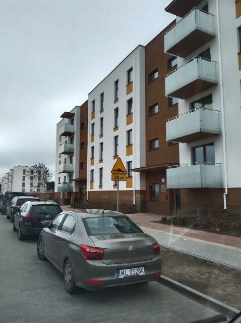 Nowoczesny budynek wielorodzinny z balkonami i zaparkowanymi samochodami wzdłuż nowej drogi z kostki brukowej. Uwagę zwraca znak drogowy ostrzegawczy.