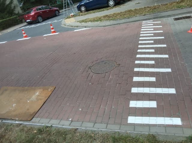 Ceglana nawierzchnia z wymalowanym przejściem dla pieszych, zabezpieczona stożkami i metalową płytą. Widoczny właz kanalizacyjny i fragment ulicy z samochodami.
