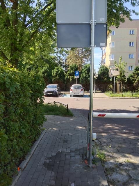 Ulica z chodnikiem z kostki, szlabanem i znakiem parkingu. W tle budynek mieszkalny i zaparkowane samochody. Widok na infrastrukturę drogową w słoneczny dzień.