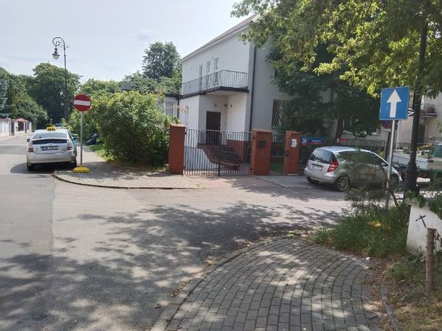 Ulica z kostki brukowej, znak zakazu wjazdu, budynek z balkonem i zaparkowane samochody. Widok na fragment drogi i otoczenie miejskie w słoneczny dzień.