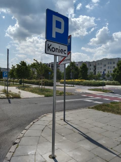 Znak drogowy z symbolem parkingu i napisem 'Koniec' na tle miejskiego krajobrazu z budynkami i drzewami. Widoczne przejście dla pieszych i fragment chodnika.