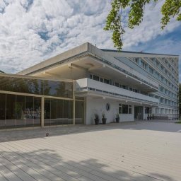 RESORT NADMORSKIE HOTELE SPÓŁKA Z OGRANICZONĄ ODPOWIEDZIALNOŚCIĄ - Elegancki budynek z przeszkloną werandą i tarasem, idealny na event. Jasna kolorystyka, nowoczesna architektura, zieleń w tle. Widok z zewnątrz, słoneczny dzień.