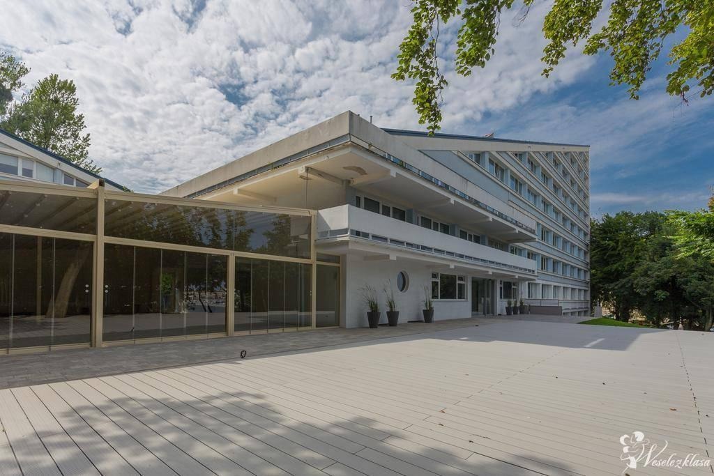 Elegancki budynek z przeszkloną werandą i tarasem, idealny na event. Jasna kolorystyka, nowoczesna architektura, zieleń w tle. Widok z zewnątrz, słoneczny dzień.