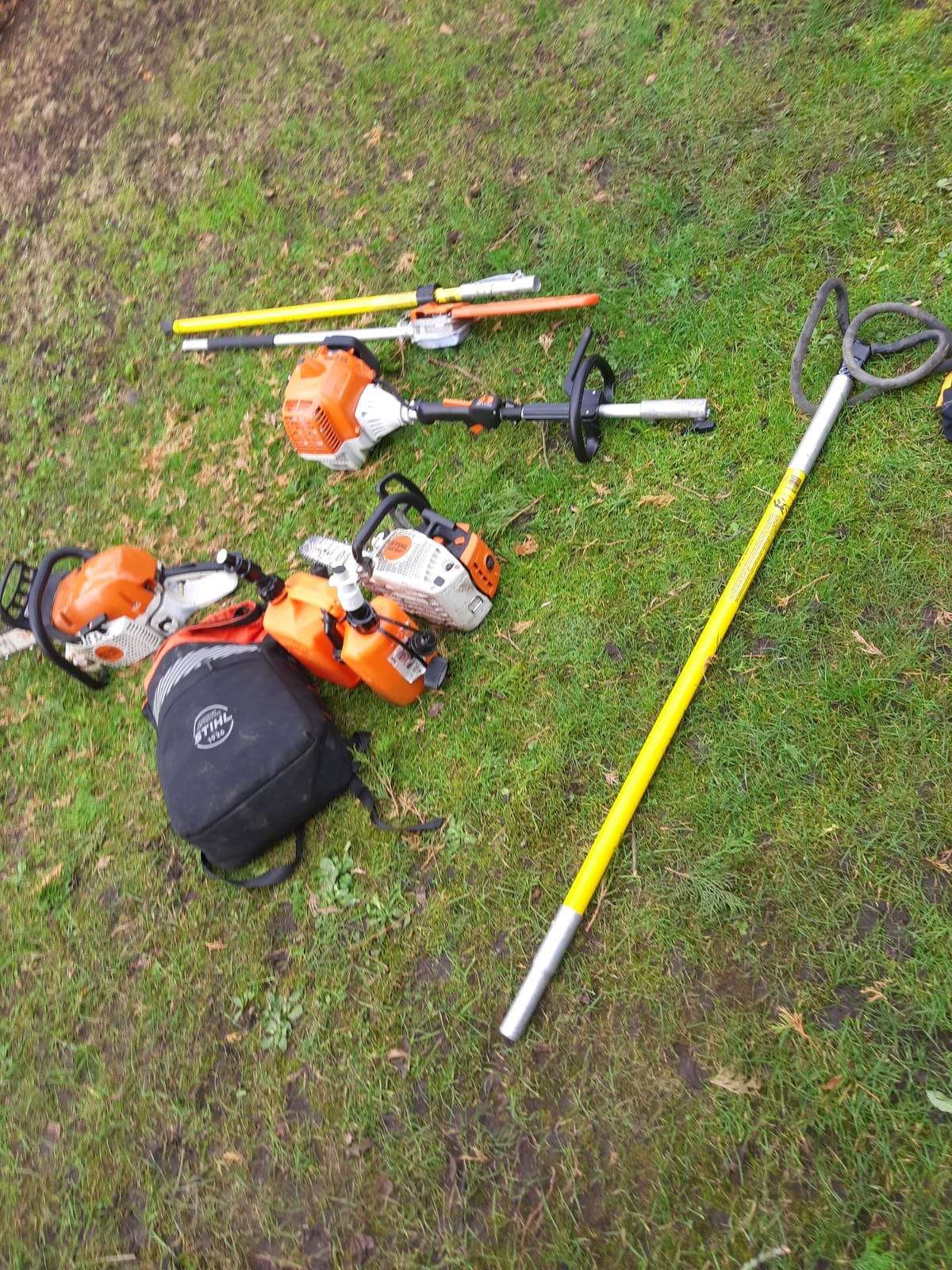 Sprzęt ogrodniczy Stihl na trawie: piły łańcuchowe, nożyce do żywopłotu na wysięgniku, podkrzesywarka i plecak. Widok z góry, narzędzia gotowe do pracy.