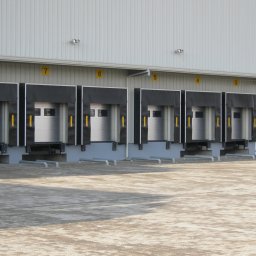 SOLID OKNA - Rząd częściowo otwartych, numerowanych bram przemysłowych z czarnymi uszczelnieniami, widocznych z zewnątrz, na tle białej hali.